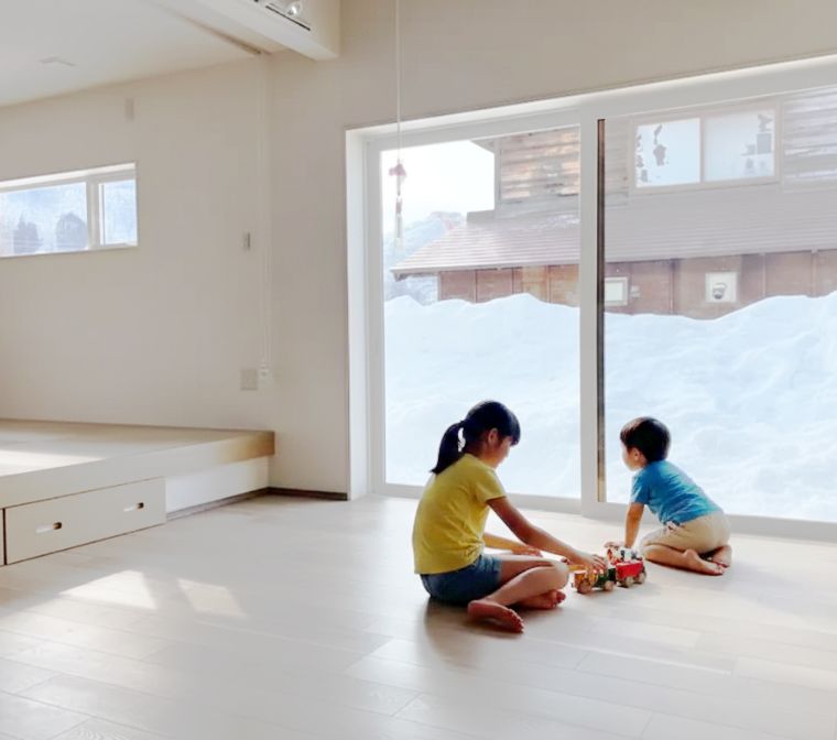 雪景色が広がる窓のそばで、子供たちが床で楽しそうに遊ぶ、明るく暖かな室内の様子