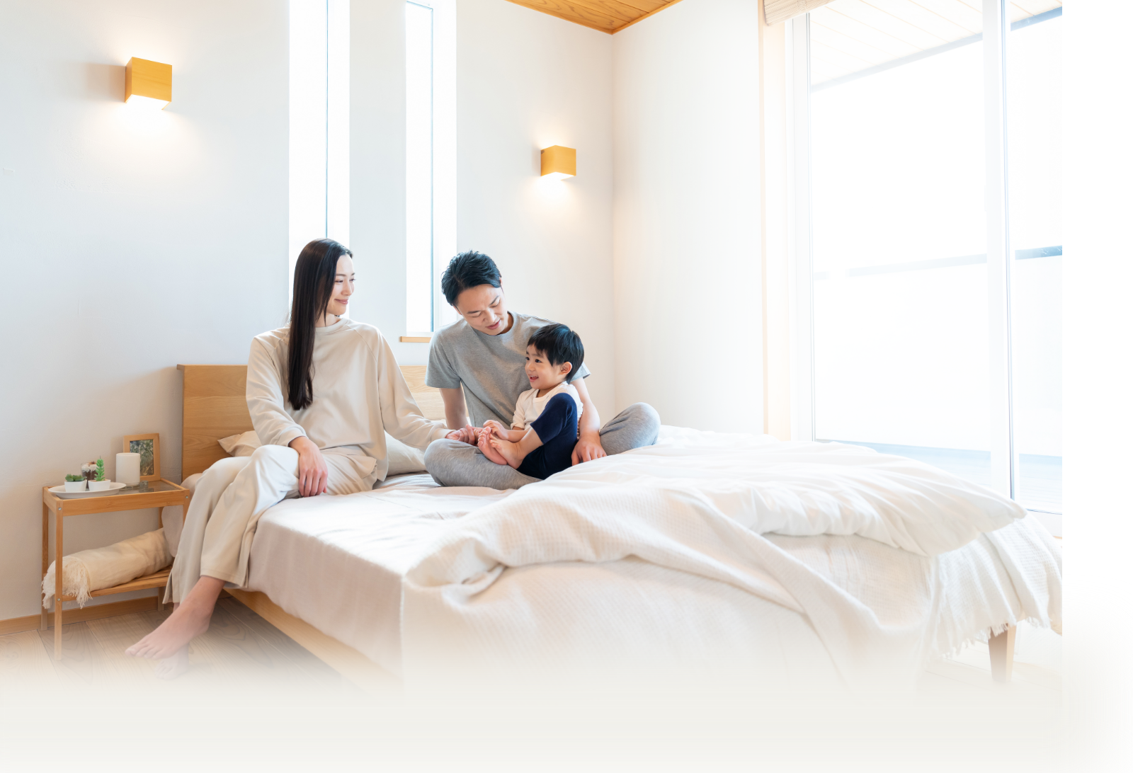 日差しが差し込む明るい寝室のベッドで、幼い息子を挟んで笑顔でくつろぐ家族の様子