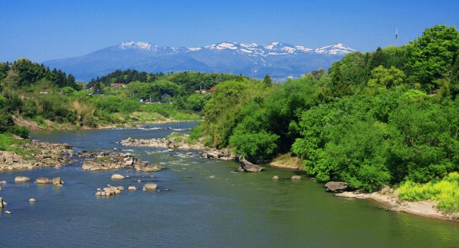 流れる川を中心に、両岸を緑の木々が覆い、遠景に雪をかぶった山々が連なる自然風景の様子