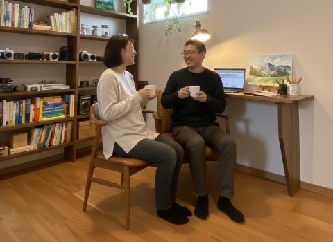 木製の書棚とデスクがある落ち着いた室内で、マグカップを手に向かい合って話す夫婦の様子