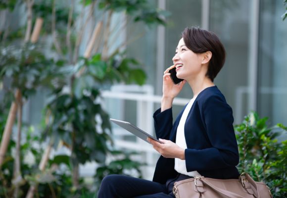 都市部の緑豊かな一角で、笑顔で電話をしながらタブレットを操作するビジネスウーマンの様子