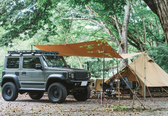 緑豊かな森の中で、オフロード車にオレンジのタープを張り、テントを設営したキャンプの様子