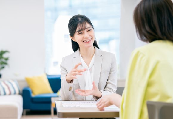 窓から光が差し込む明るい室内で、若い女性がお客様に笑顔で身振りを交えながら説明している様子