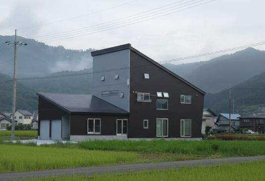水田が広がる田園地帯に、遠く霞む山々を背景に、傾斜のある屋根が特徴のモダンな家が建つ様子
