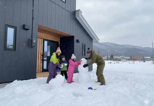 雪が降り積もった庭先で、ダークグレーのモダンな家の前を背景に、親子4人が雪だるまを作って楽しんでいる様子