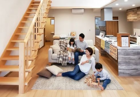 木製の階段がある明るいリビングで、父親は娘と本を読み、母親は床でマグカップ片手にくつろぎ、息子は木のブロックで遊んでいる家族4人の様子