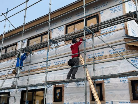 建設中の2階建て住宅の外壁工事で、足場の上で二人の作業員が断熱材や下地を取り付けている様子
