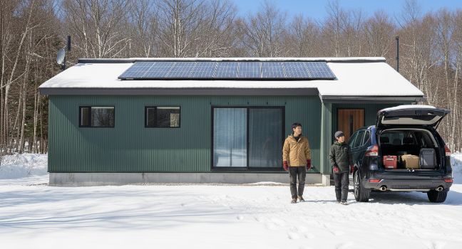 雪に覆われた景色の中、ソーラーパネルが載る緑色の家の前で、荷物を積んだSUVと2人が立つ様子