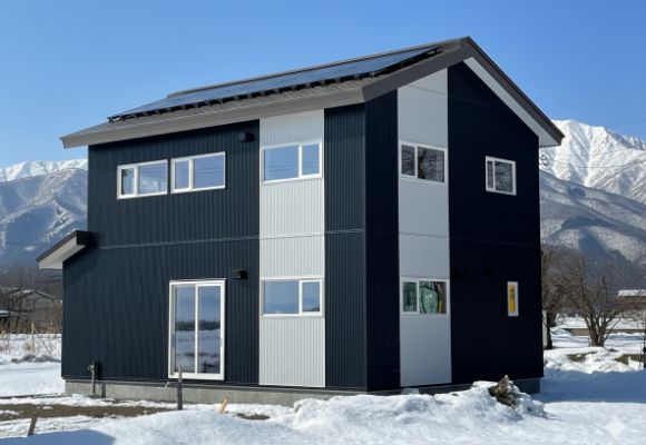雪景色が広がる中に建つ、ダークブルーとグレーのモダンな2階建て住宅の様子。屋根にはソーラーパネルがあり、背景には雪山が見える