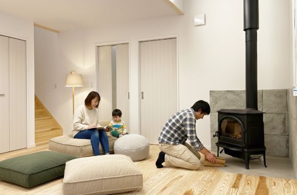 薪ストーブのあるリビングで、父親が薪をくべ、母親が子供に本を読んでくつろぐ家族の様子
