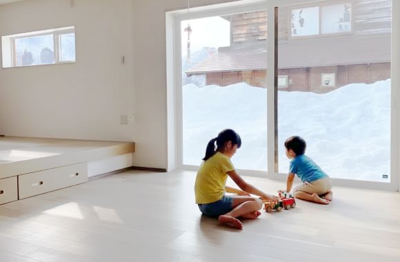 雪景色が広がる窓のそばで、子供たちが床で楽しそうに遊ぶ、明るく暖かな室内の様子